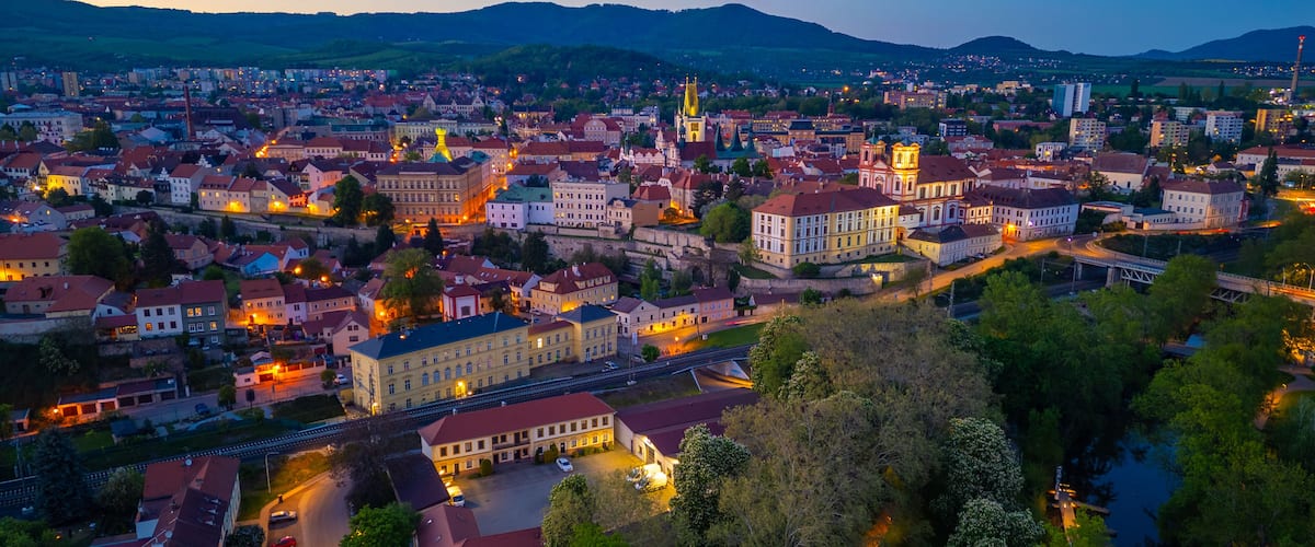 Una vista panoramica di una città al crepuscolo, con edifici illuminati, un campanile di una chiesa in evidenza e un fiume sullo sfondo.
