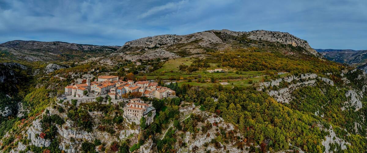 A village perched on a hillside with terracotta-roofed buildings, surrounded by rugged mountains and autumn foliage.
