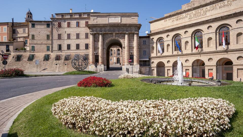 A historic building with an ornate archway, a fountain, and a flower bed.