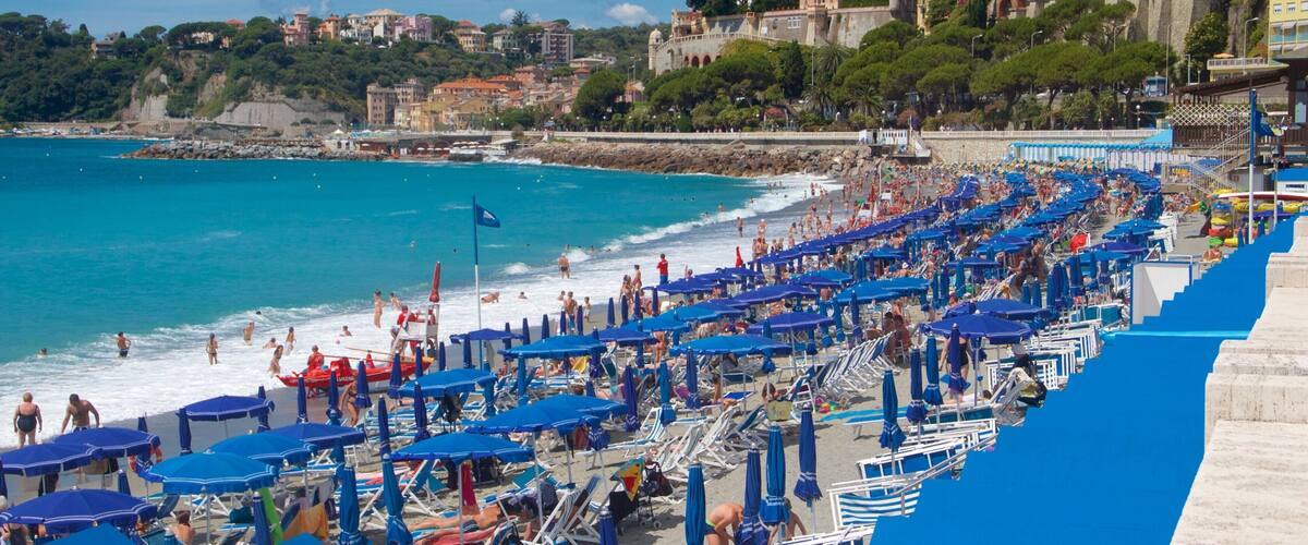 A crowded beach with blue umbrellas and people enjoying the water.