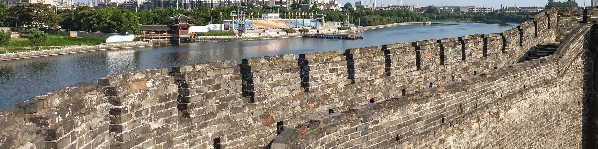Un muro de piedra histórico junto a un río, con edificios modernos al fondo.