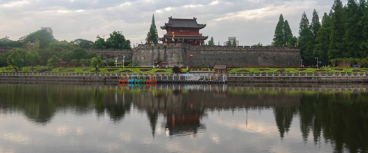 A traditional Chinese building with a reflection in the water, surrounded by greenery and a stone wall.