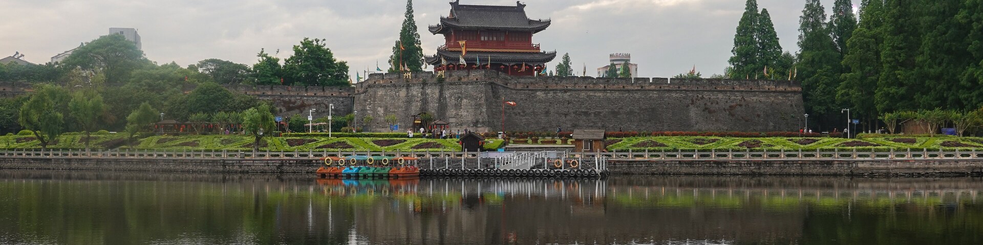A traditional Chinese building with a reflection in the water, surrounded by greenery and a stone wall.