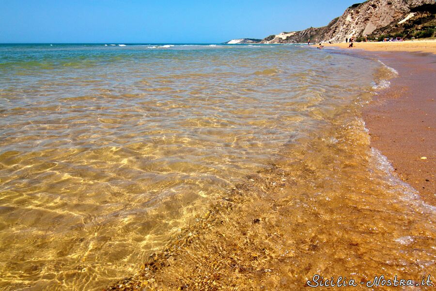 A beach with clear shallow waters, a sandy shore, and rocky cliffs in the background.