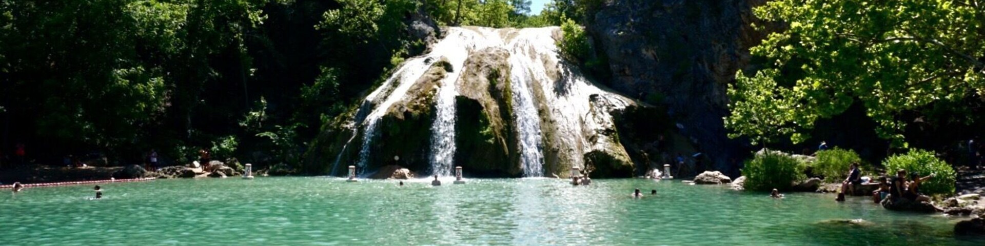 Uma cascata deságua em uma piscina turquesa límpida, rodeada por vegetação exuberante e paredões rochosos.