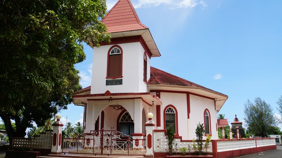 A church with a red roof and white walls, surrounded by trees and a fence.