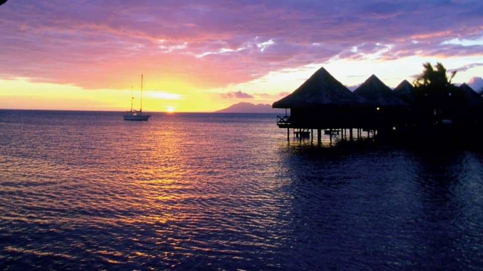 A sunset over the ocean with a sailboat, palm trees, and overwater bungalows.