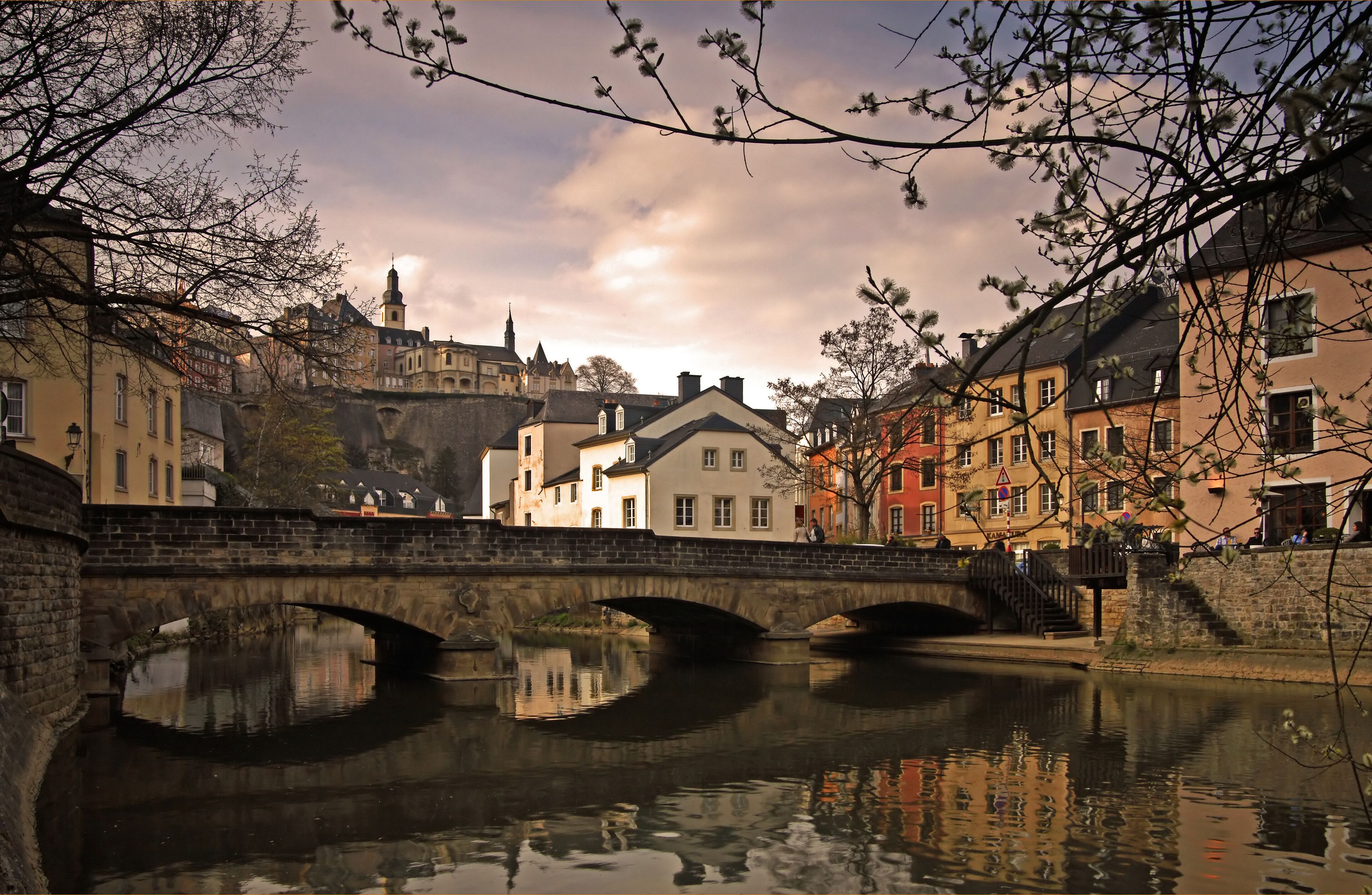 A stone bridge over a calm river, reflecting buildings and a castle in the background.