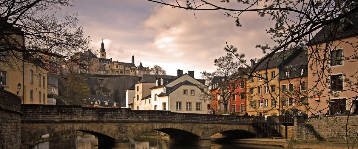 A stone bridge over a calm river, reflecting buildings and a castle in the background.