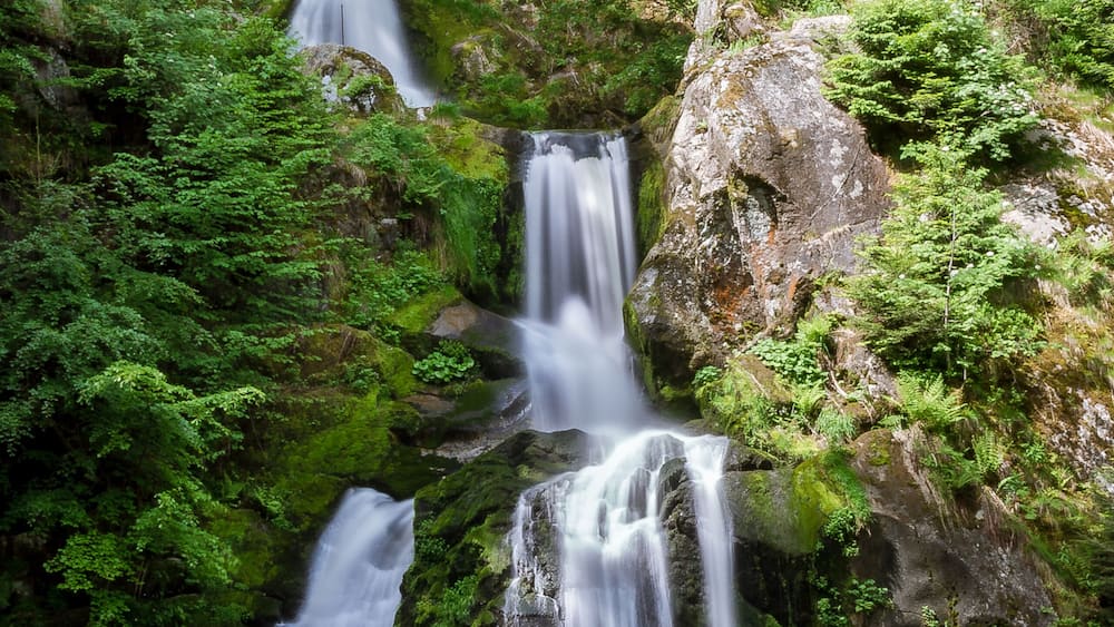 Ein mehrstufiger Wasserfall, umgeben von ĂŒppigem GrĂŒn und einer HolzbrĂŒcke.