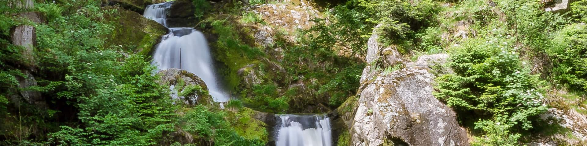 A multi-tiered waterfall surrounded by lush greenery and a wooden bridge.