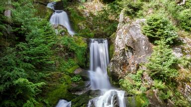 Uma cascata com vários níveis, cercada por vegetação exuberante e uma ponte de madeira.
