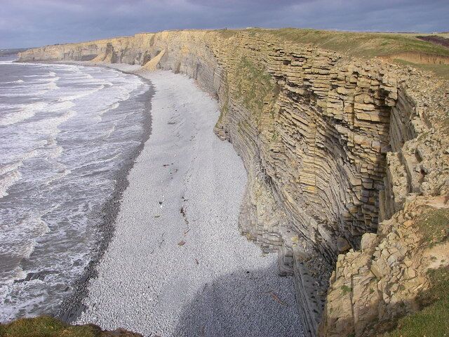 A coastal cliff with layered sedimentary rock formations and a pebbly beach.