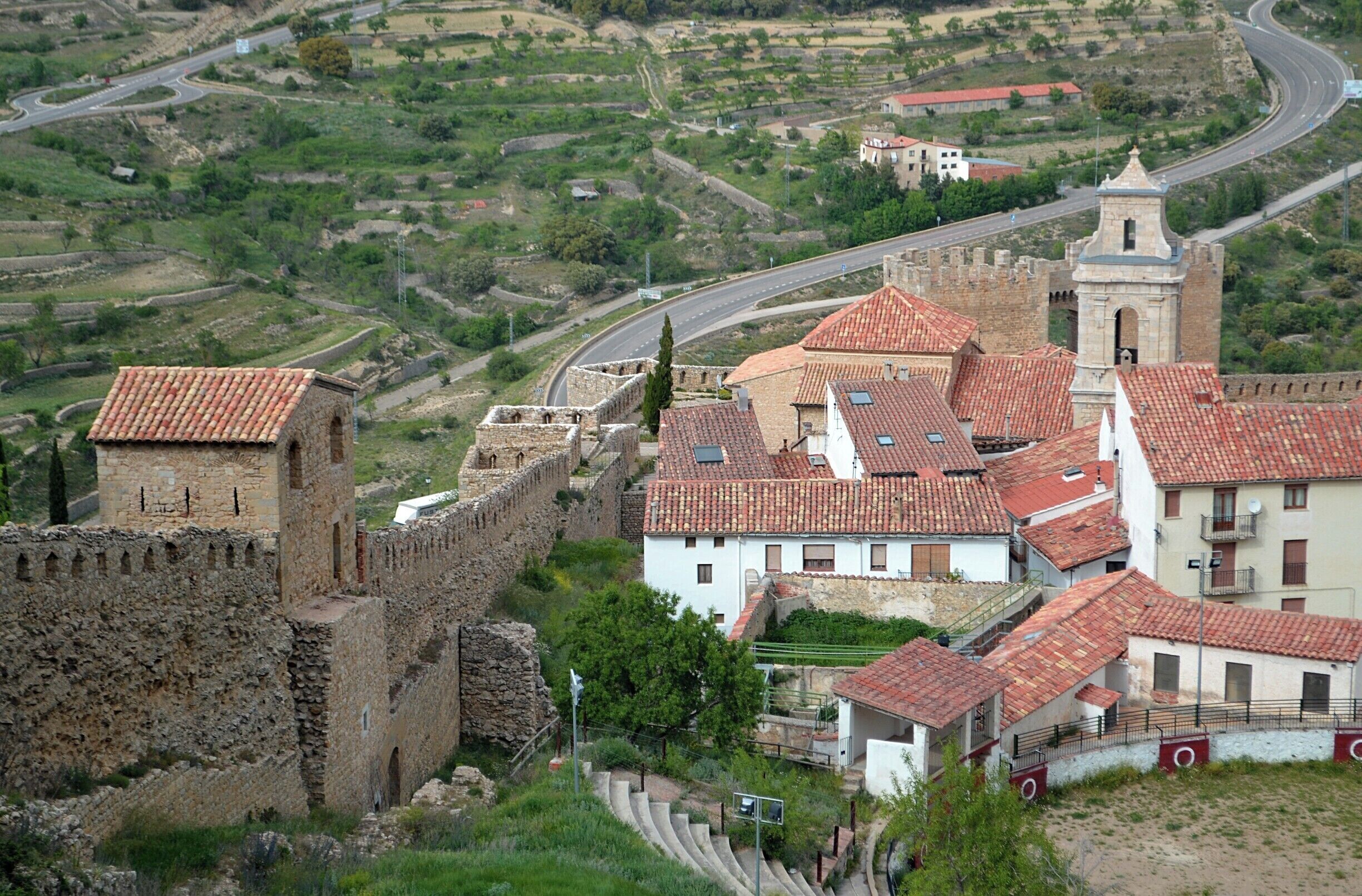 A historic town with a fortified wall, a church with a bell tower, and terraced fields in the background.