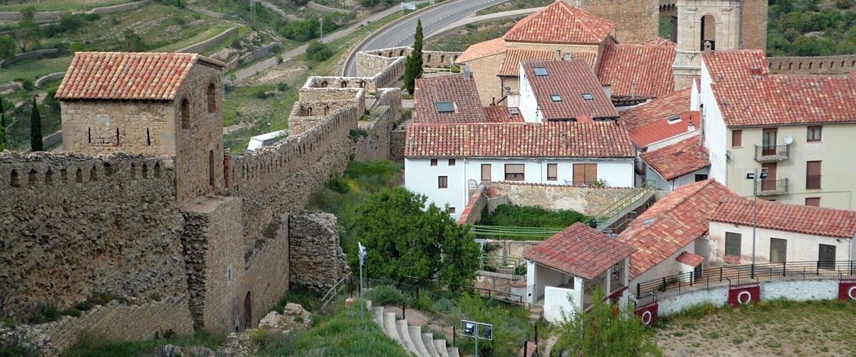 A historic town with a fortified wall, a church with a bell tower, and terraced fields in the background.