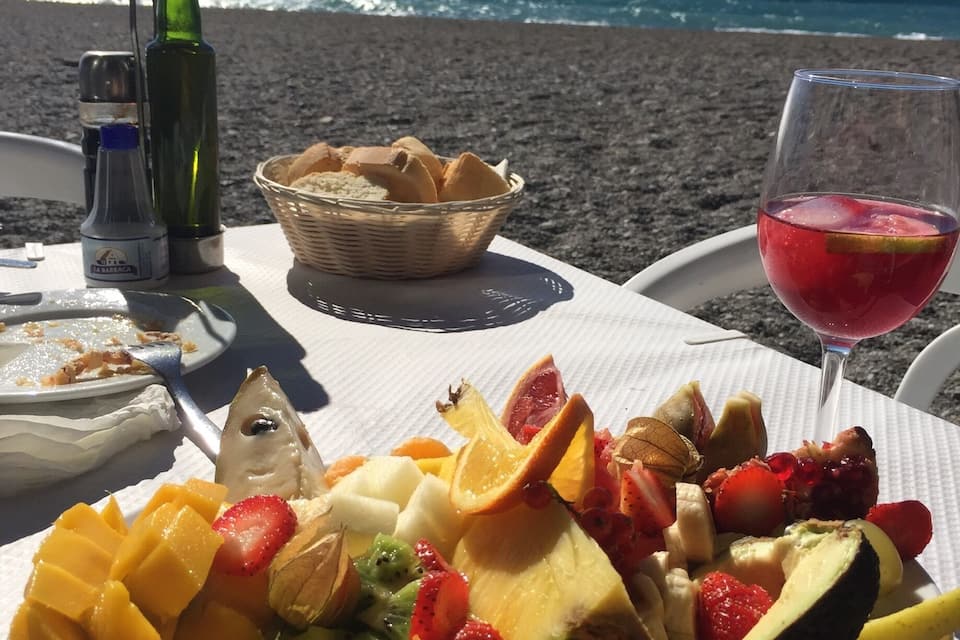 A table with a fruit salad, bread, and a glass of red wine on a beach.