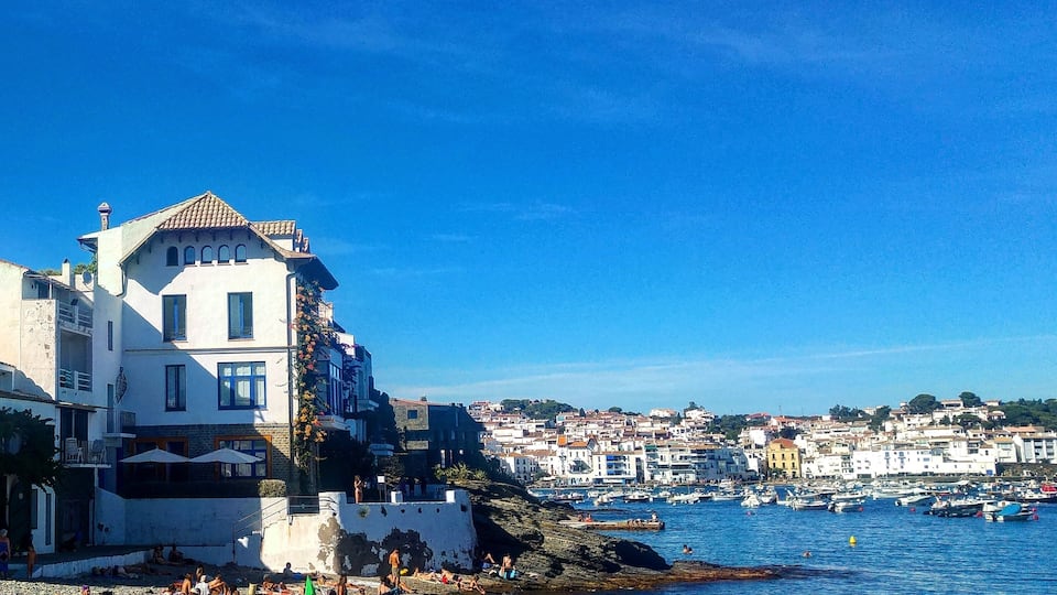 A beach with people sunbathing and swimming, a rocky shoreline, and a town in the background.