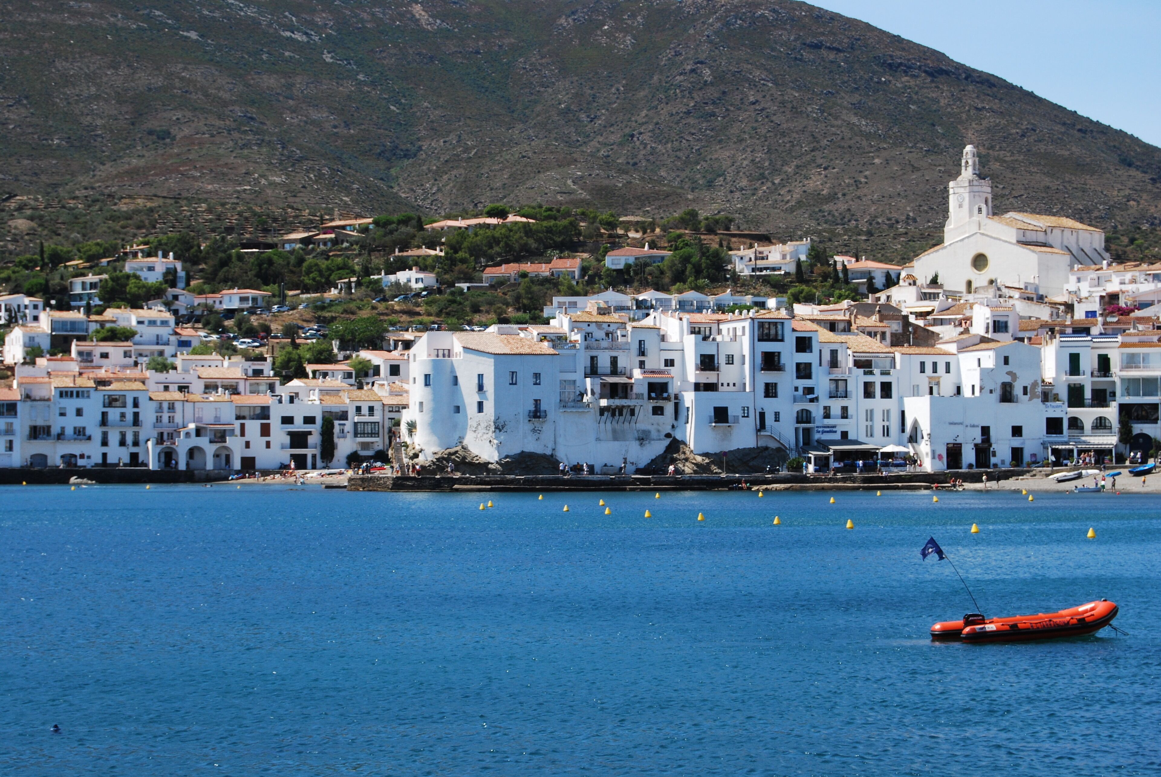 A coastal town with white buildings, a church, and a mountain in the background.