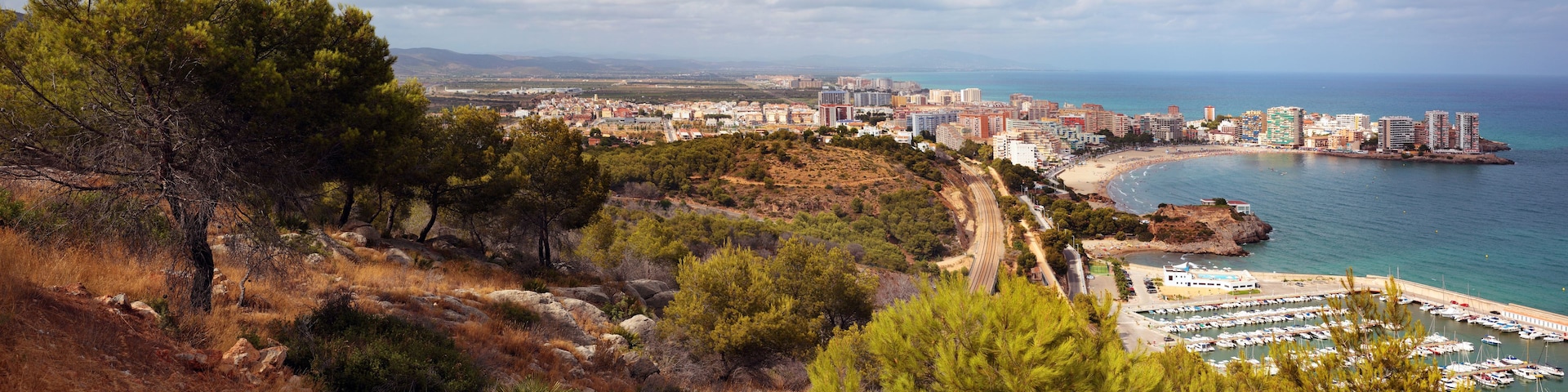 A coastal cityscape with a beach, marina, and mountainous terrain.