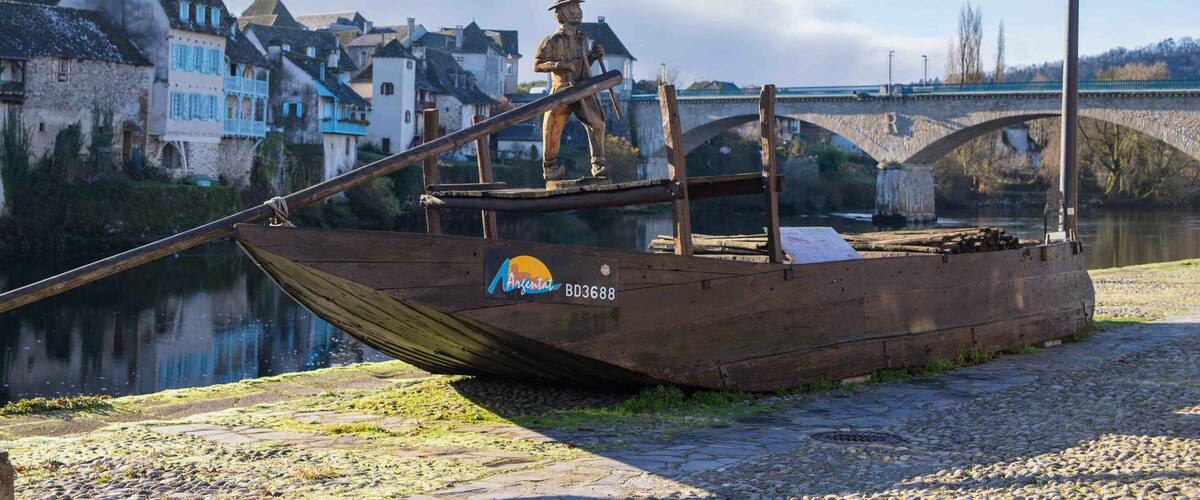 A wooden boat with a statue on it, a bridge in the background, and houses along the riverbank.