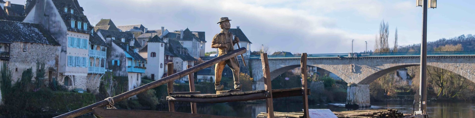 A wooden boat with a statue on it, a bridge in the background, and houses along the riverbank.