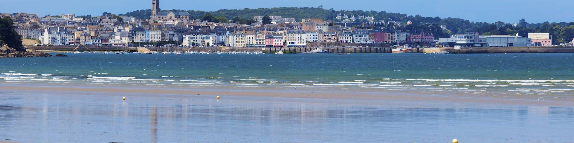 A coastal town with a prominent church tower, a sandy beach, and a harbor with boats.