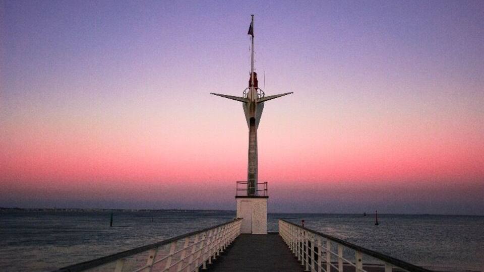 A pier leading to a tall tower with a flag at sunset.