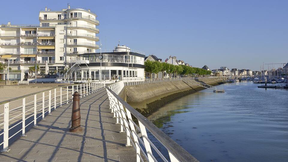 A waterfront promenade with a white railing, a large building with balconies, and a calm river with boats.