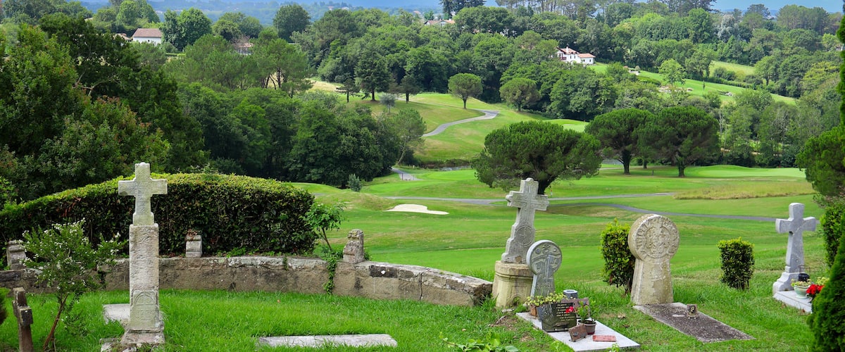 A cemetery with stone crosses and headstones, surrounded by lush greenery and a golf course in the background.