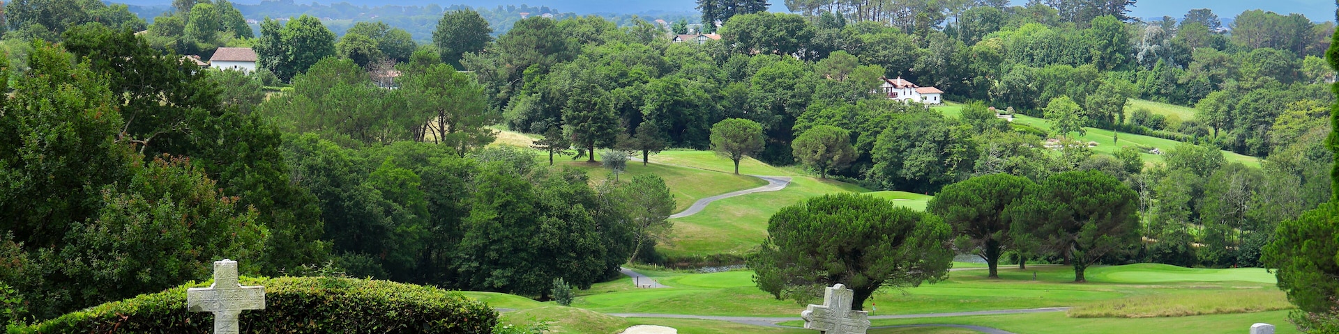 A cemetery with stone crosses and headstones, surrounded by lush greenery and a golf course in the background.