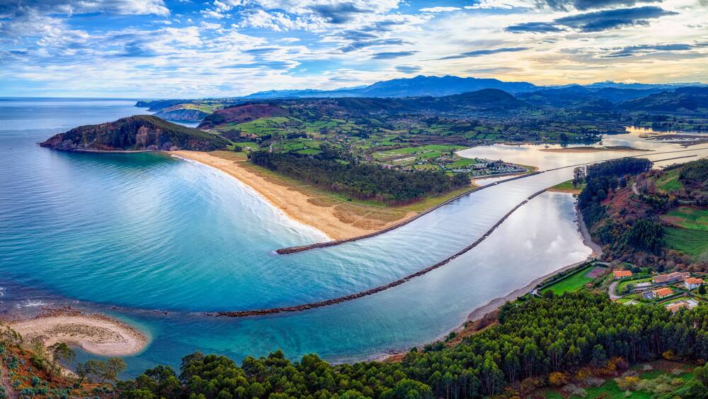 A coastal landscape with a beach, a calm sea, a river, and distant mountains.