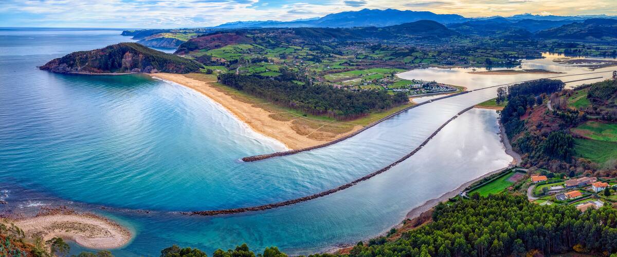 A coastal landscape with a beach, a calm sea, a river, and distant mountains.
