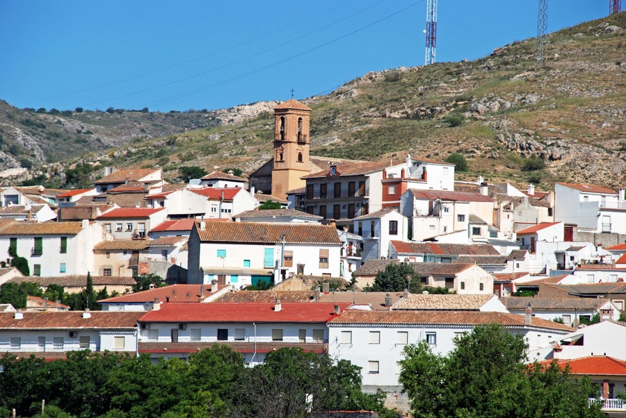 Une ville perchée sur une colline, avec un clocher d’église imposant, des bâtiments blancs aux toits rouges et un ciel d’un bleu éclatant.