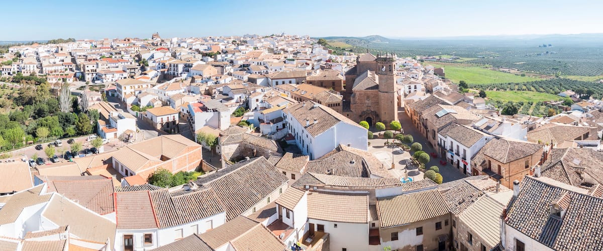 A town with terracotta-roofed buildings, a prominent church with a bell tower, and a hilly landscape in the background.