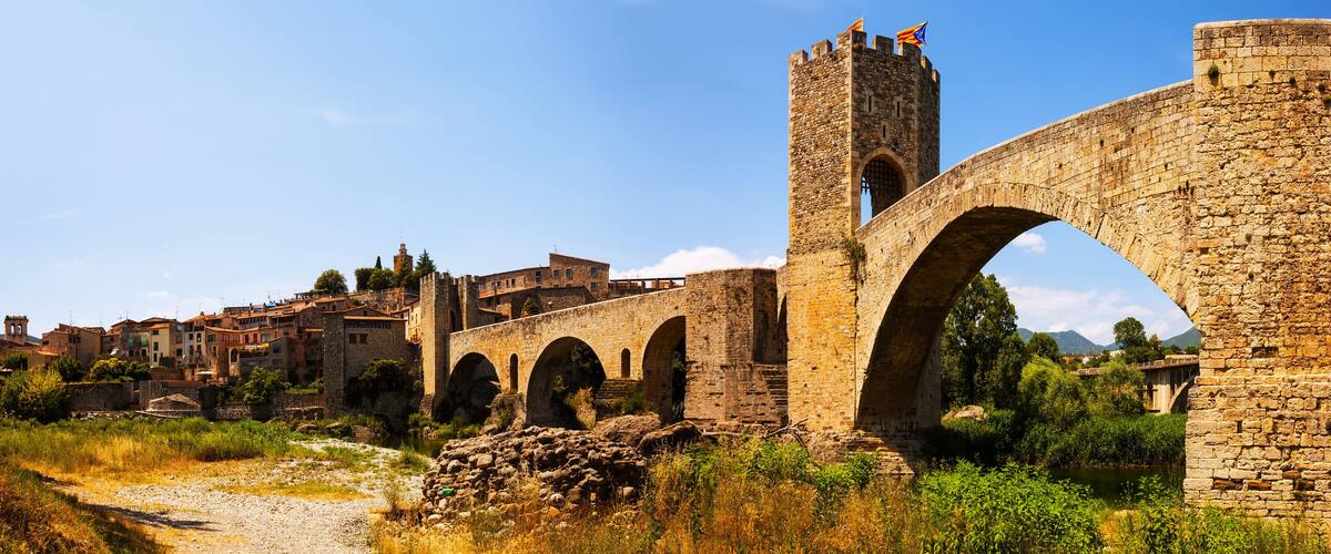 An ancient stone bridge with multiple arches, a tower, and historic buildings in the background.