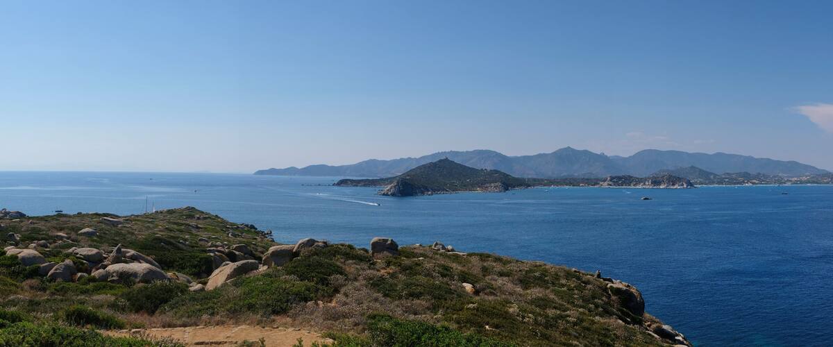A coastal landscape with rocky terrain, a clear blue sea, and distant mountains.