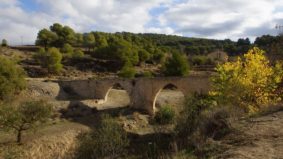 A stone bridge over a dry riverbed, surrounded by sparse vegetation and a backdrop of hills with trees.