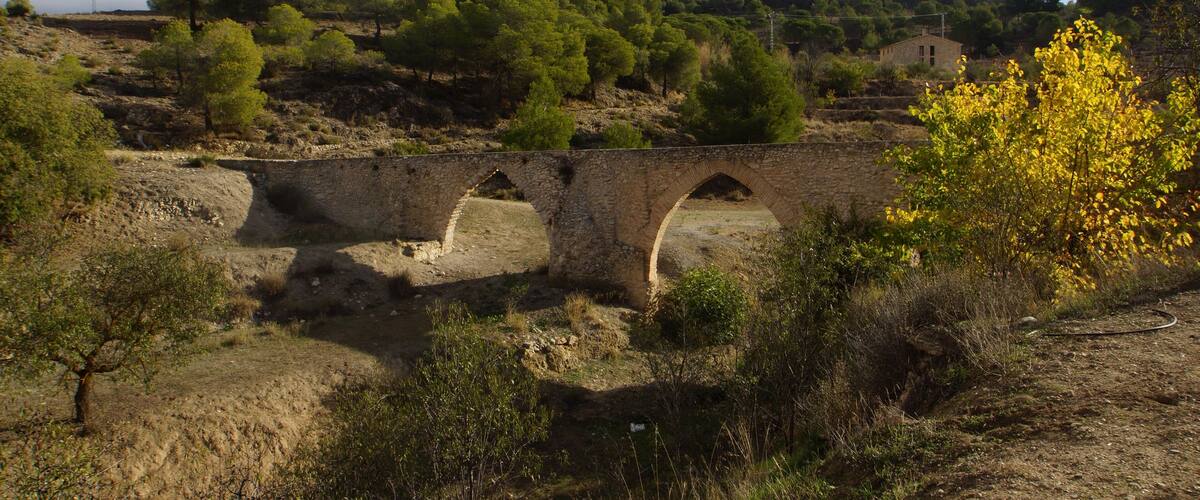 A stone bridge over a dry riverbed, surrounded by sparse vegetation and a backdrop of hills with trees.