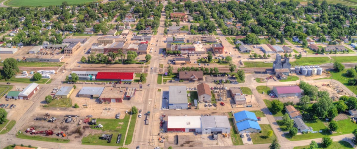 An aerial view of a town with a central road, surrounding buildings, and green fields.