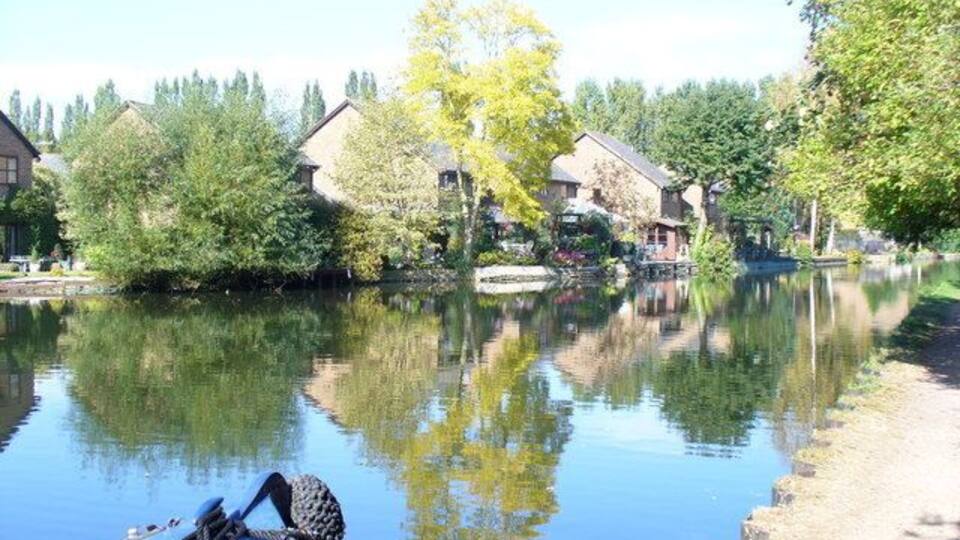 A calm river with houses on the opposite bank, a boat in the foreground, and trees reflecting on the water.