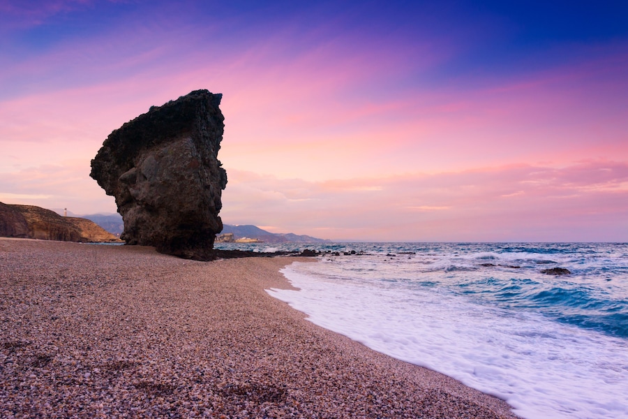 Uma praia de seixos com uma grande formação rochosa e o oceano.