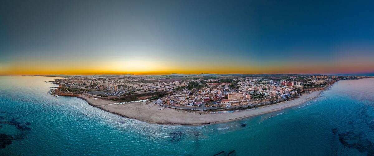 An aerial view of a coastal town with a beach, buildings, and a sunset over the ocean.