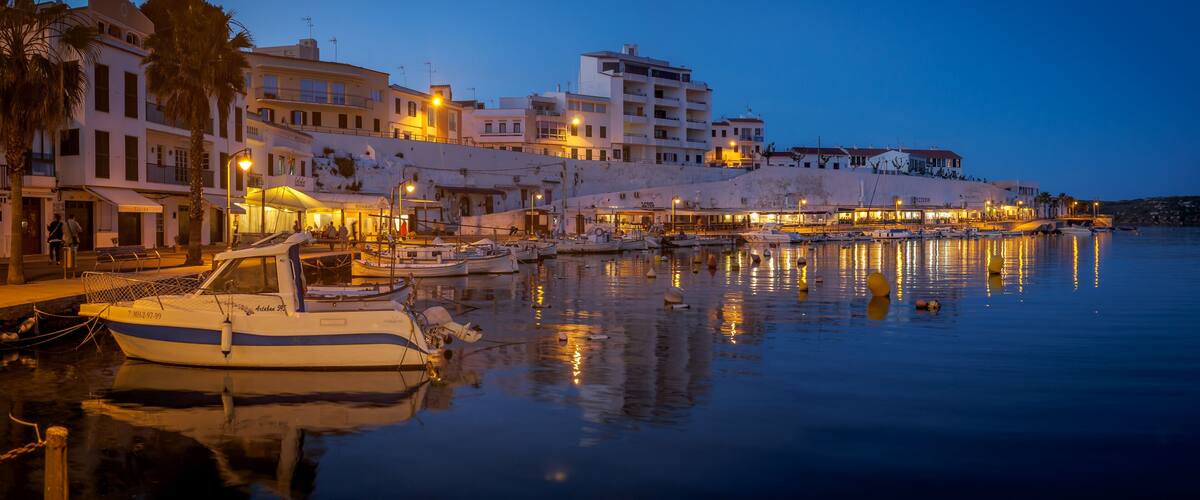 A marina at dusk with boats docked, waterfront buildings, and a calm sea reflecting the lights.