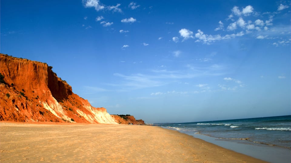 A beach with a red cliff, clear sky, and calm sea.