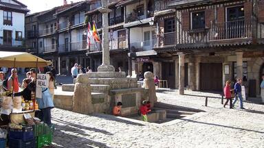 Uma praça de calçada com uma fonte, bancas de mercado e edifícios tradicionais.