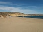 Ein Sandstrand mit einer Holztreppe, die zum Wasser führt, einige Personen und hügeliges, felsiges Gelände im Hintergrund.
