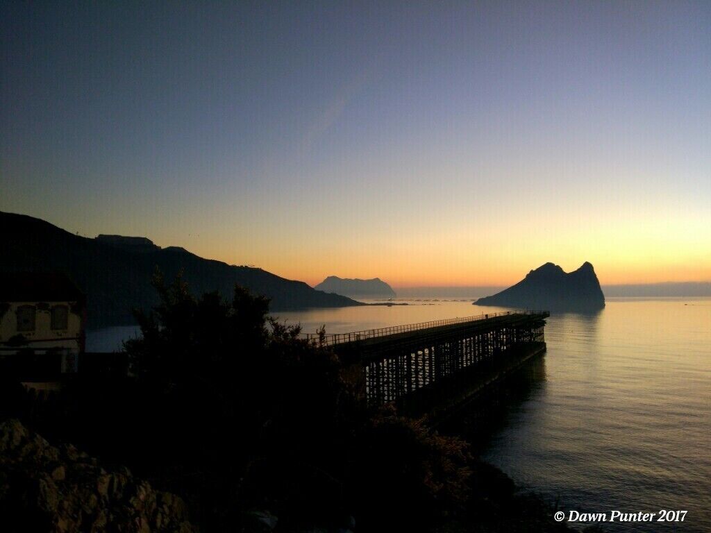 A sunset over a calm sea with a pier extending into the water, surrounded by mountains.
