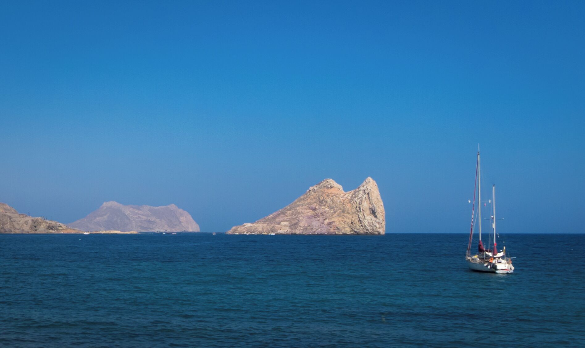 A sailboat on calm blue waters with rocky islands in the background.