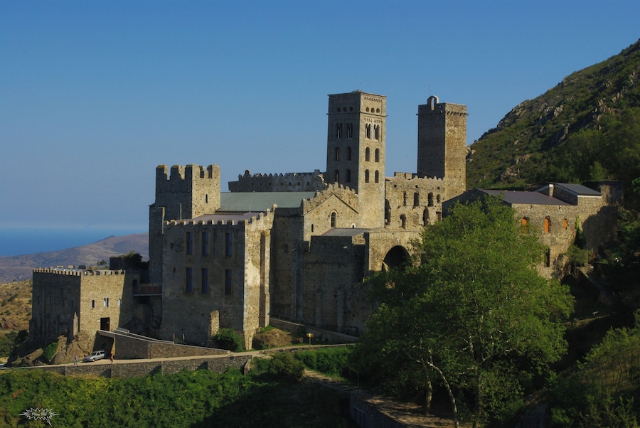 A historic castle with multiple towers and a fortified wall, surrounded by lush greenery and hills.