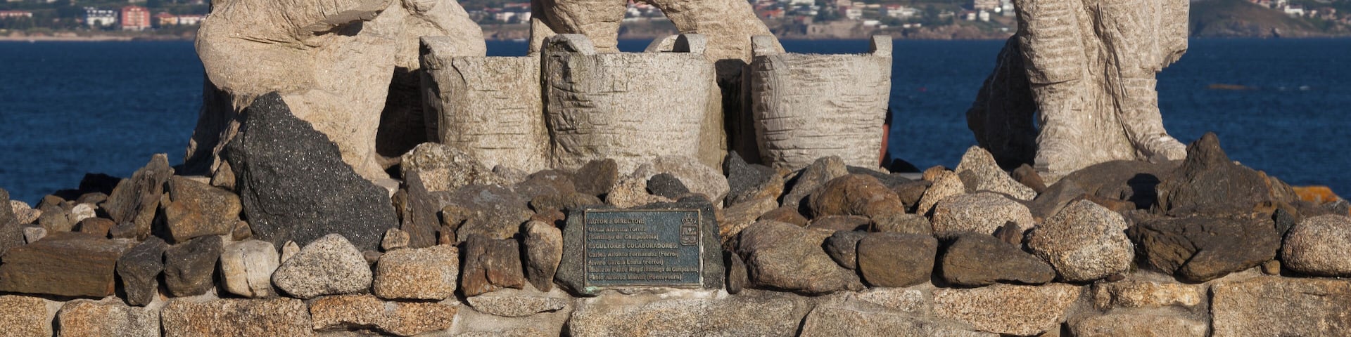 Three stone statues on a rocky outcrop with a coastal town in the background.